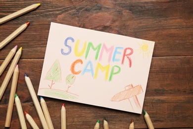 Photo of Card with text SUMMER CAMP, drawings and colorful pencils on wooden table, flat lay