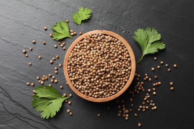 Photo of Dried coriander seeds in bowl and green leaves on dark gray textured table, flat lay