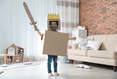 Photo of Cute little boy playing cardboard armor in living room