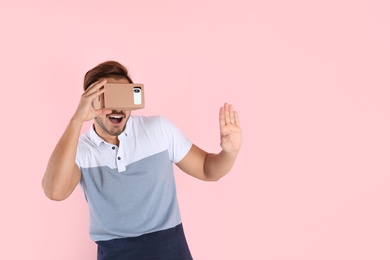 Young man using cardboard virtual reality headset on color background. Space for text