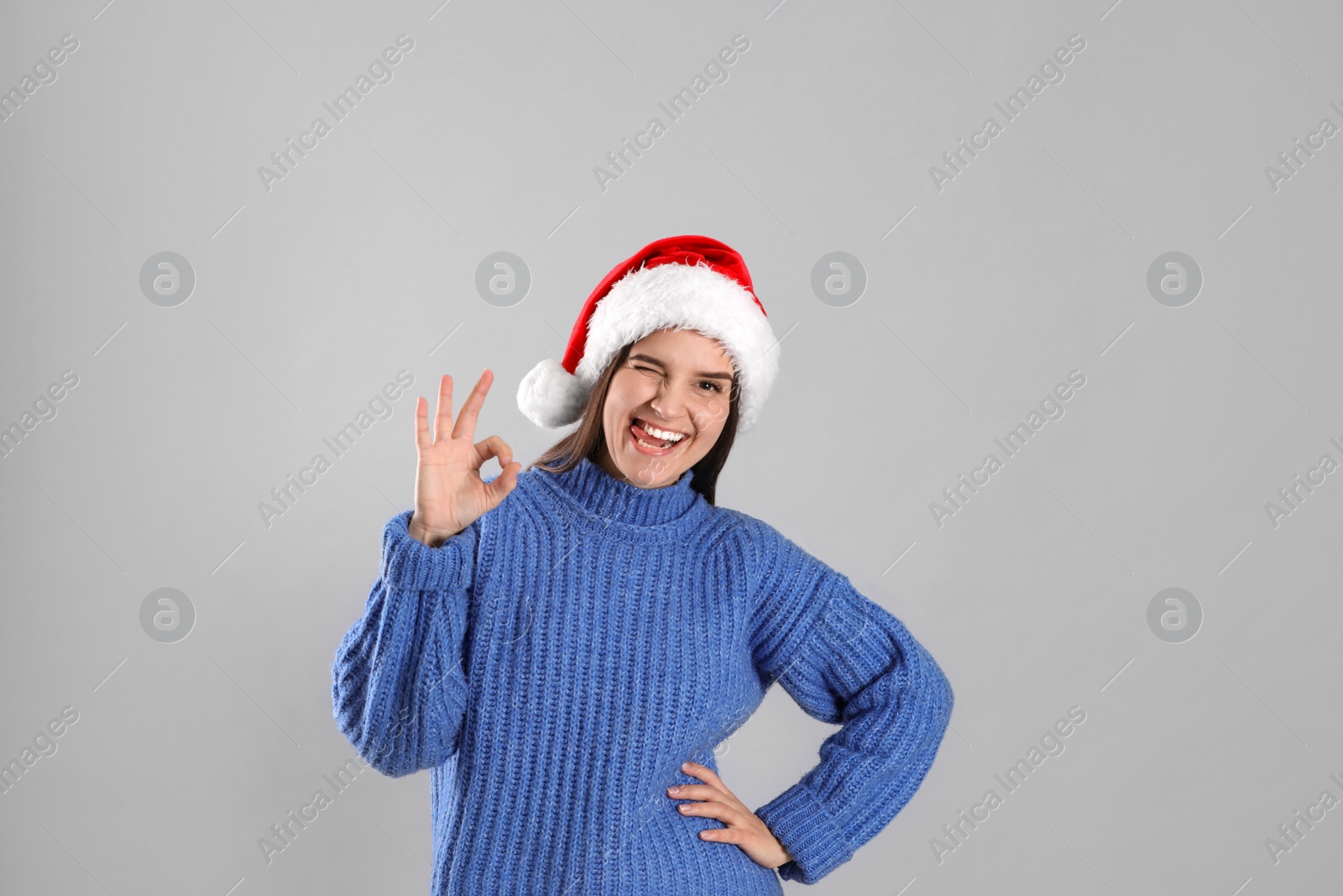 Photo of Pretty woman in Santa hat and blue sweater showing OK gesture on grey background