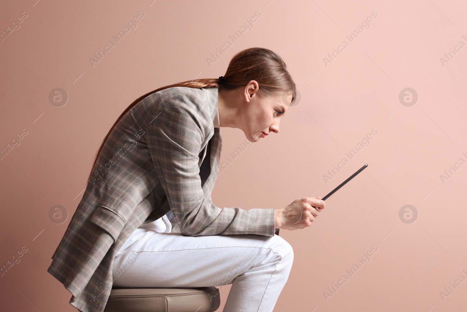 Photo of Woman with bad posture using tablet while sitting on stool against pale pink background