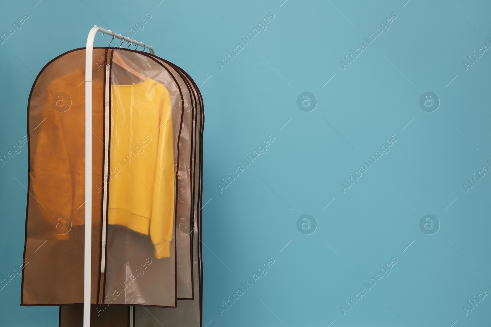 Photo of Garment bags with clothes on rack against light blue background. Space for text