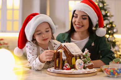 Photo of Mother and daughter decorating gingerbread house at table indoors