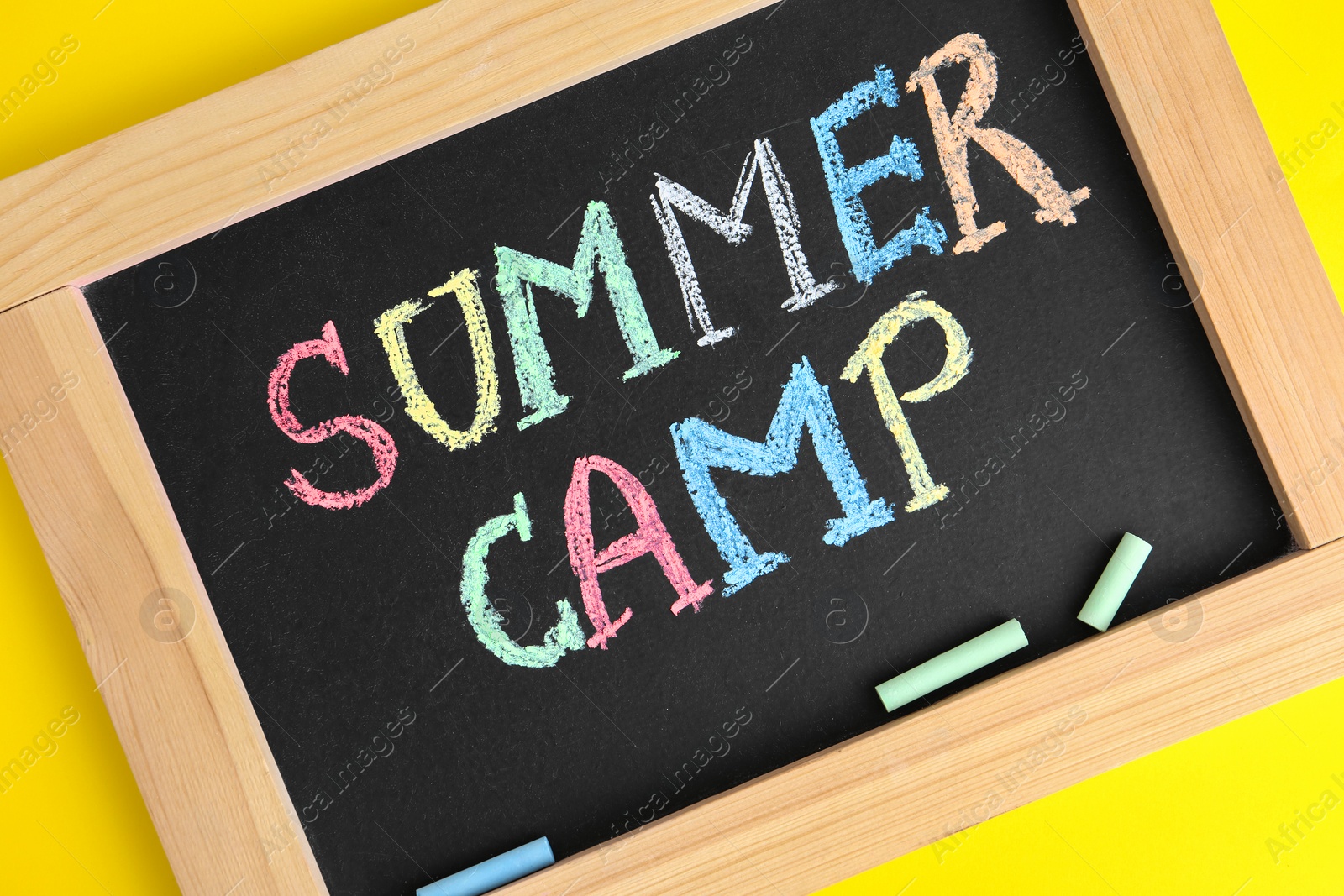 Photo of Blackboard with words SUMMER CAMP and colorful chalk on yellow background, closeup