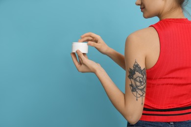 Woman with tattoo holding jar of cream against light blue background, closeup. Space for text