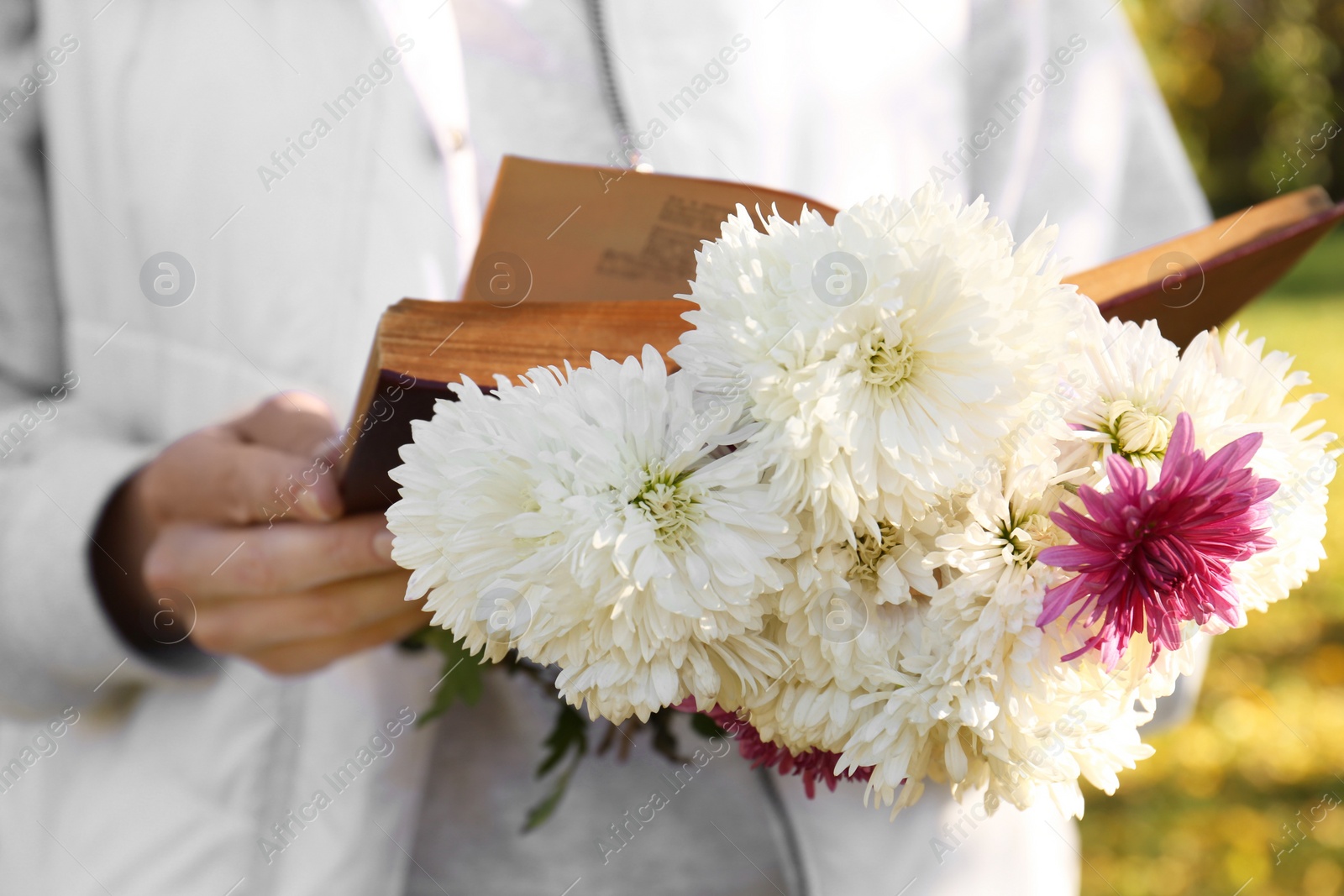 Photo of Woman reading book and holding beautiful flowers outdoors on autumn day, closeup