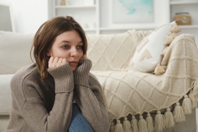 Sad young woman sitting near sofa at home, space for text