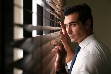 Photo of Handsome young man looking through window blinds indoors