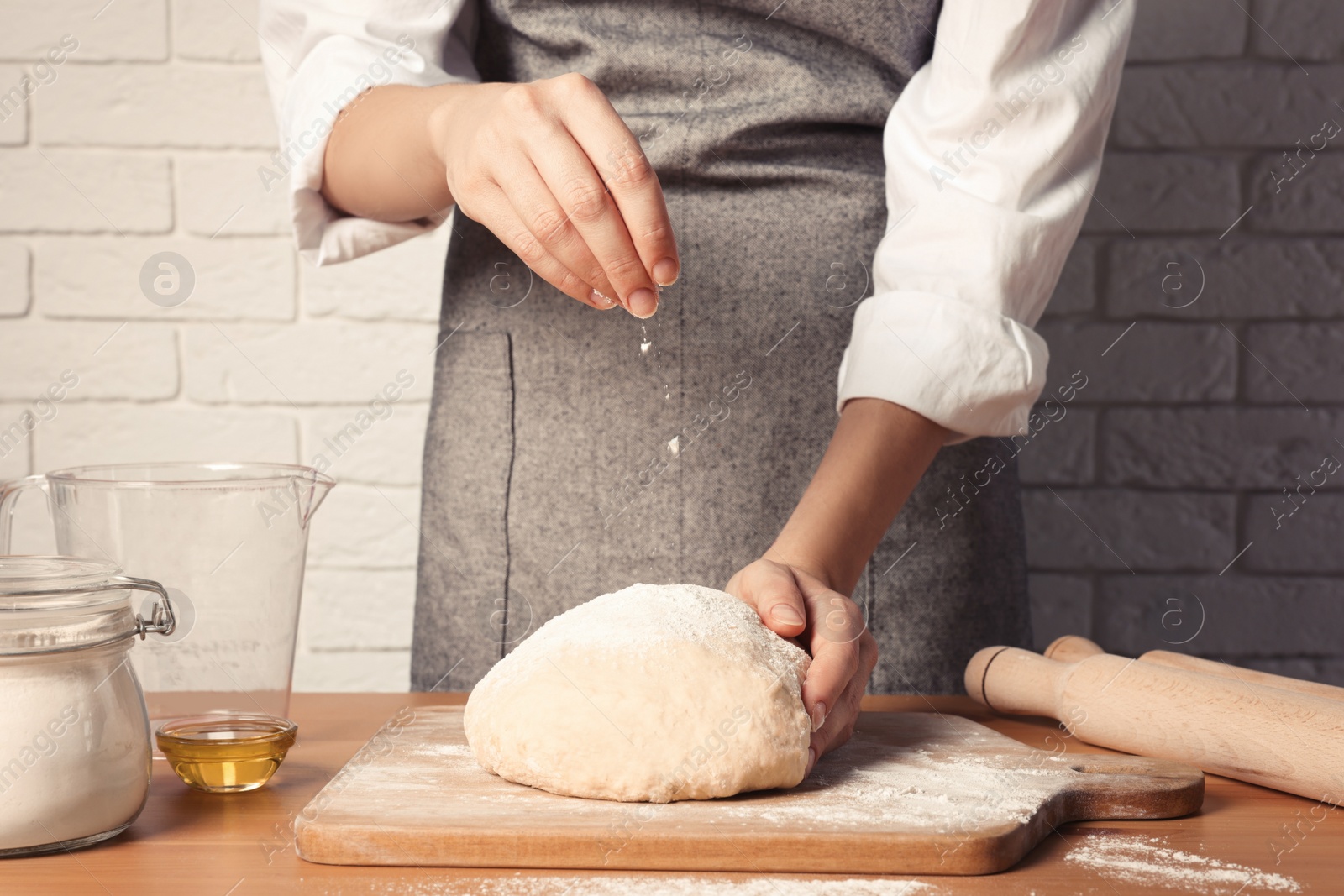 Photo of Woman sprinkling flour over dough at table near white brick wall, closeup