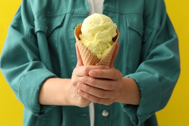 Woman holding tasty ice cream in wafer cone on yellow background, closeup