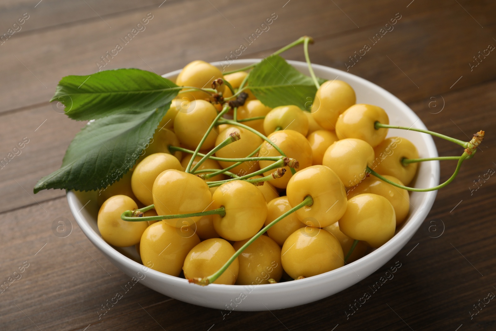 Photo of Bowl with ripe yellow cherries and green leaves on wooden table, closeup