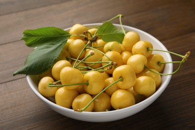 Photo of Bowl with ripe yellow cherries and green leaves on wooden table, closeup