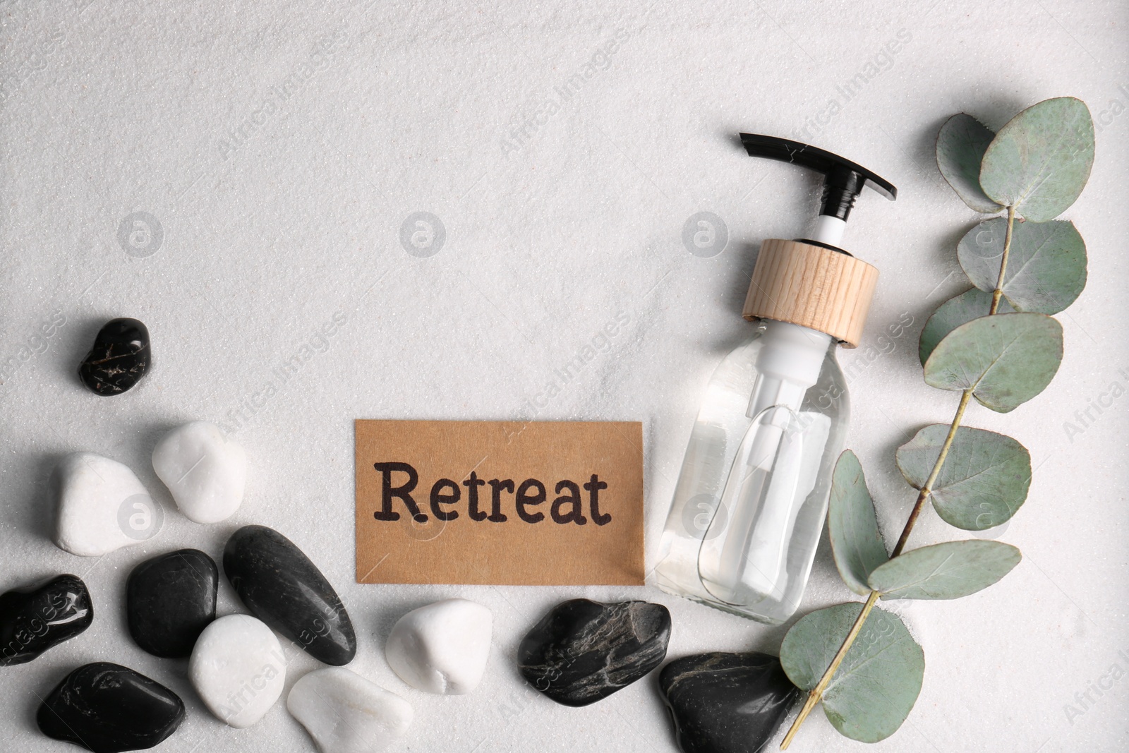 Photo of Card with word Retreat, eucalyptus branch, cosmetic bottle and stones on white sand, flat lay. Space for text