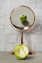 Photo of Bitten green apple near mirror with reflection of whole fruit on table