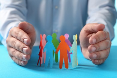 Photo of Man protecting paper human figures on light blue table, closeup. Diversity and inclusion concept