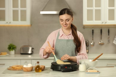 Photo of Happy woman cooking delicious crepe on electric maker at white marble table in kitchen