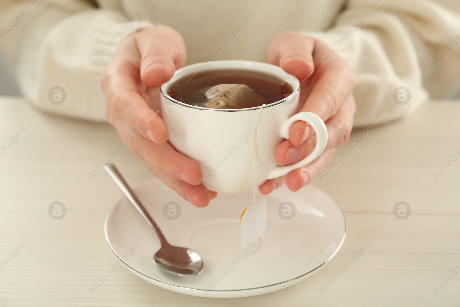 Photo of Woman holding cup with tea bag and hot water at white wooden table, closeup