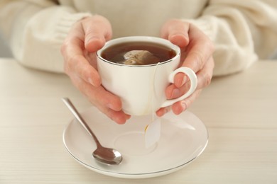 Woman holding cup with tea bag and hot water at white wooden table, closeup