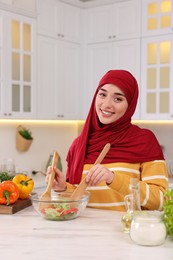 Muslim woman making delicious salad with vegetables at white table in kitchen