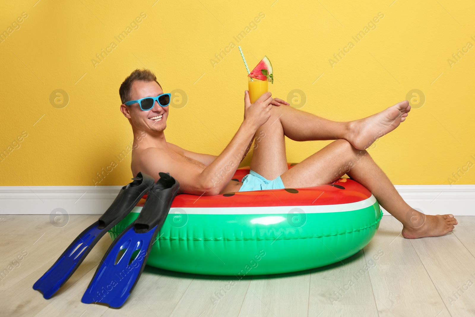 Photo of Shirtless man with inflatable ring,  flippers and glass of cocktail on floor near color wall