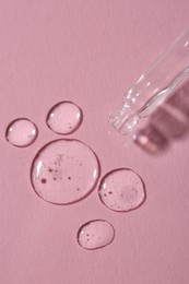 Dripping cosmetic serum from pipette onto pink background, top view
