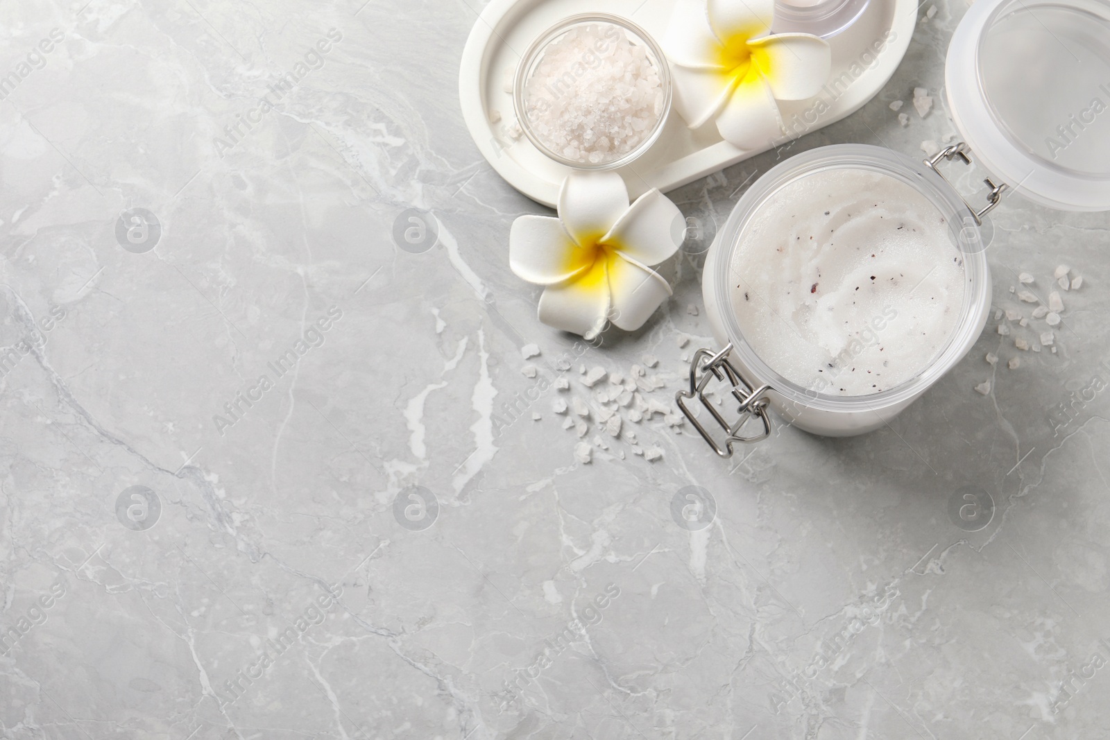 Photo of Body scrub in glass jar, plumeria flowers and sea salt on grey marble table, flat lay. Space for text