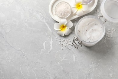 Body scrub in glass jar, plumeria flowers and sea salt on grey marble table, flat lay. Space for text