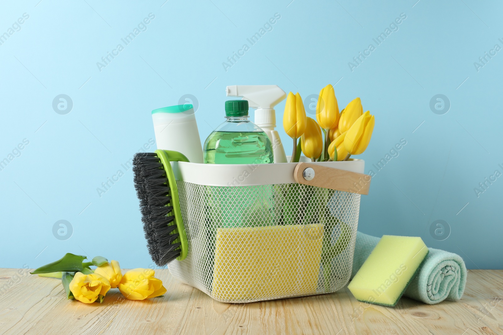 Photo of Basket with different cleaning supplies and beautiful spring flowers on wooden table against light blue background