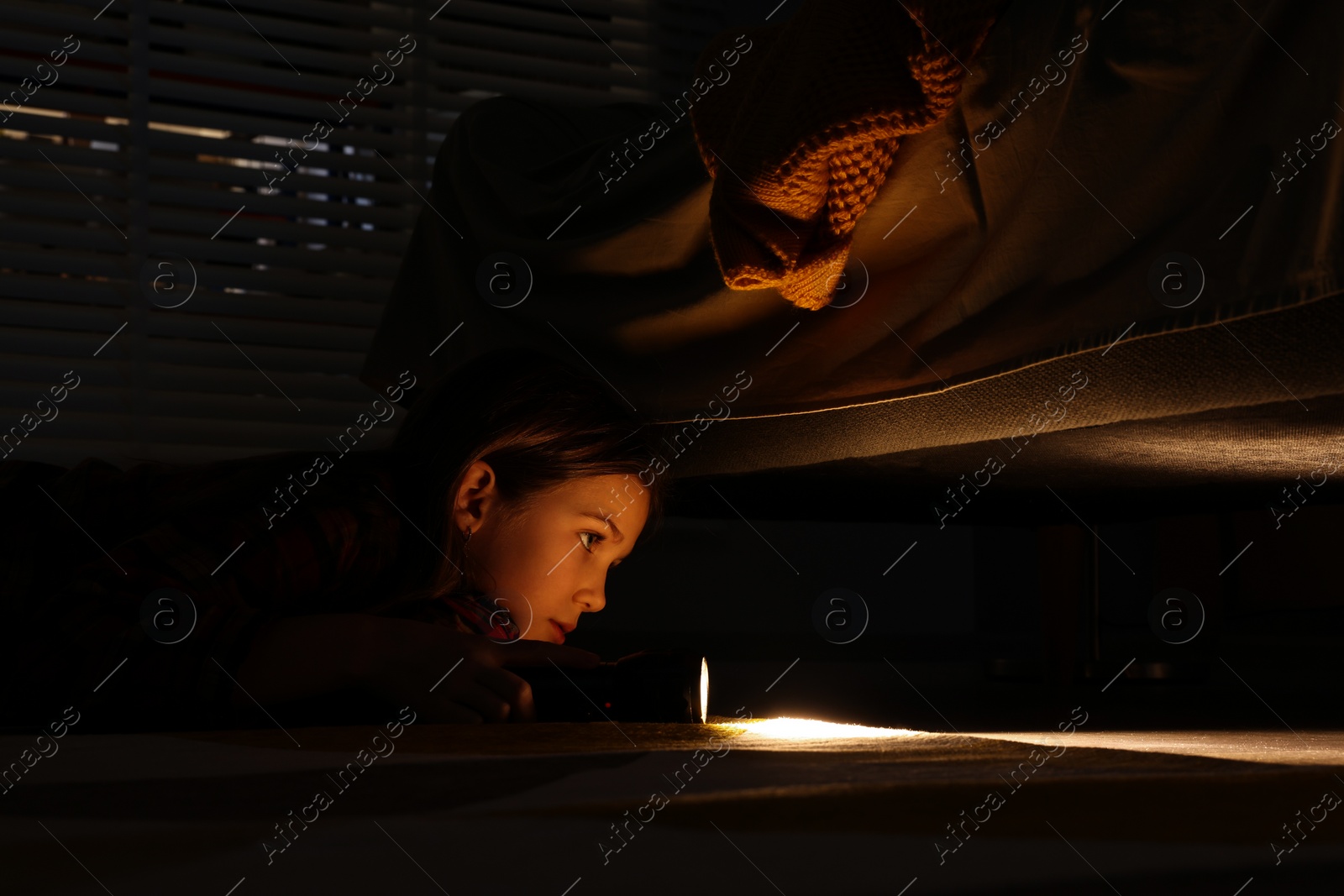 Photo of Little girl with flashlight looking for monster under bed at night