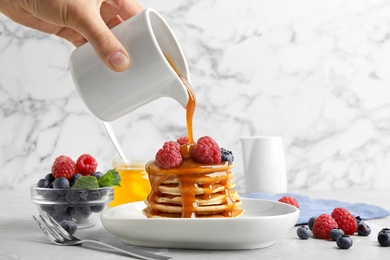 Photo of Woman pouring syrup onto fresh pancakes with berries at light grey marble table, closeup