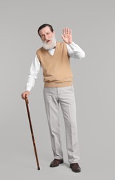 Senior man with walking cane waving on light gray background