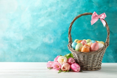 Wicker basket with painted Easter eggs and flowers on table against color background, space for text