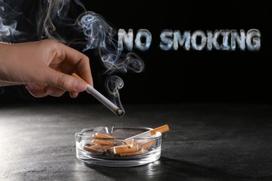 Image of No Smoking, phrase of smoke. Woman holding smoldering cigarette over glass ashtray at grey table against black background, closeup