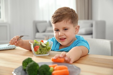 Photo of Adorable little boy eating vegetables and salad at table in room