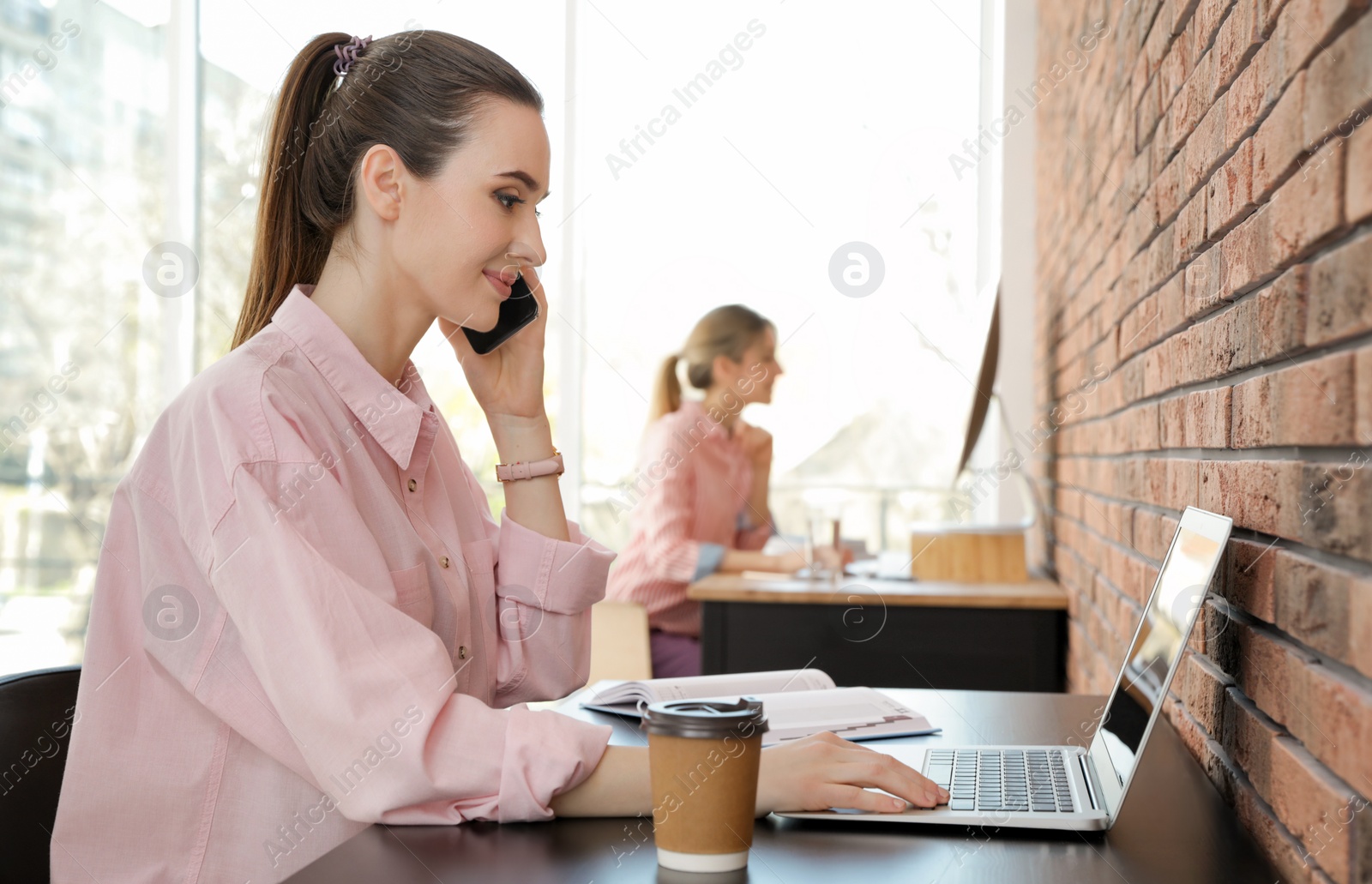 Photo of Young businesswoman talking on phone while using laptop at table in office