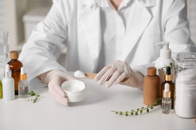 Dermatologist with jar testing cosmetic product at white table indoors, closeup