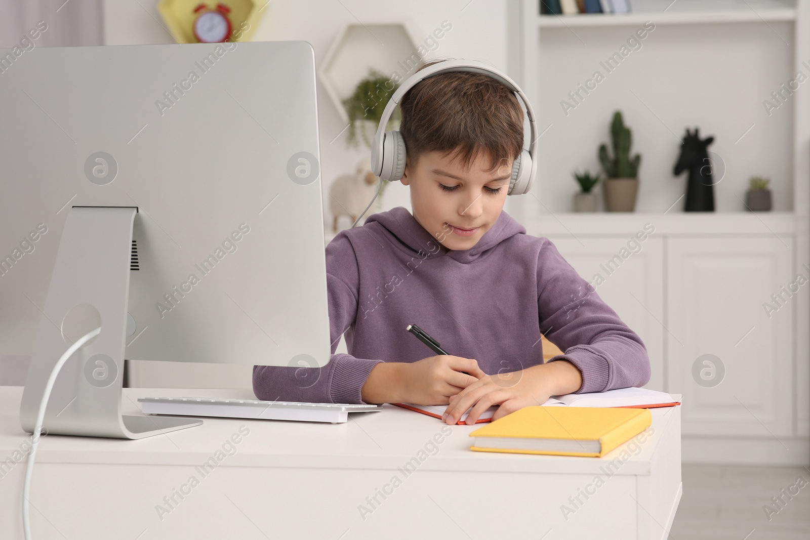 Photo of Boy writing in notepad while using computer and headphones at desk in room. Home workplace