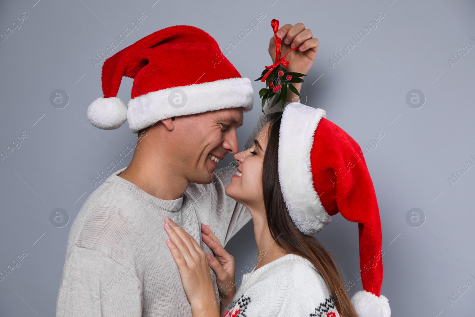 Photo of Happy couple in Santa hats under mistletoe bunch on grey background