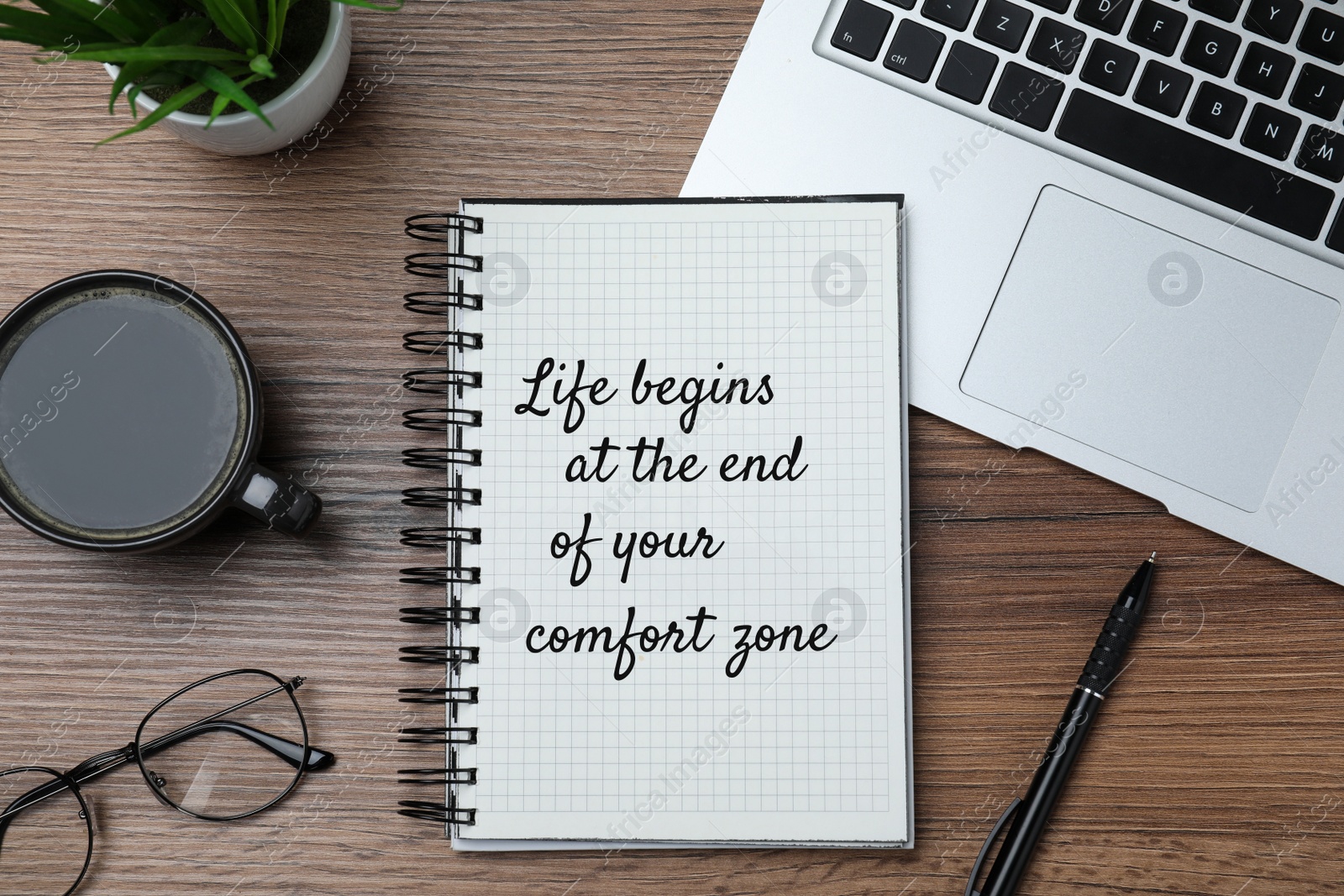 Image of Life Begins At The End Of Your Comfort Zone. Motivational quote inspiring to do something new, different from ordinary life. Text in notebook, laptop, cup of coffee, eyeglasses and houseplant on wooden table, flat lay