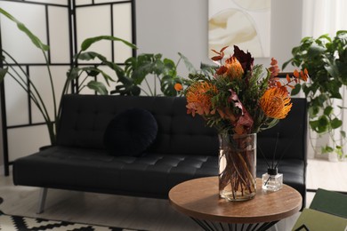 Photo of Vase with bouquet of beautiful leucospermum flowers on side table near black sofa and houseplants in room
