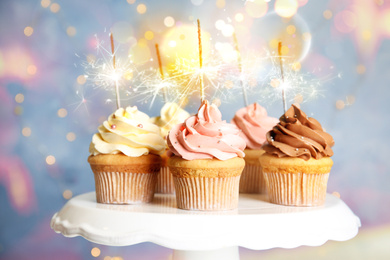 Delicious birthday cupcakes with sparklers on stand against blurred background
