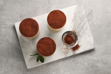 Photo of Delicious tiramisu in glasses, mint and sieve with cocoa powder on grey textured table, top view