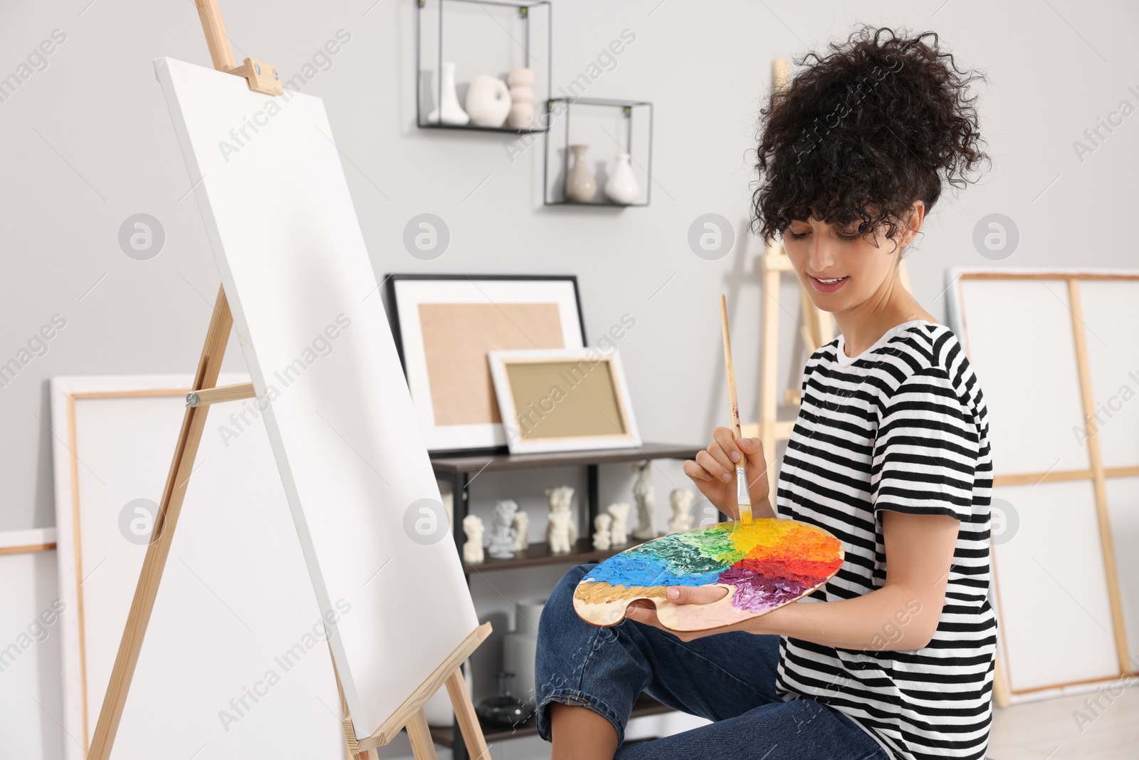 Photo of Young woman mixing paints on palette with brush near easel in studio