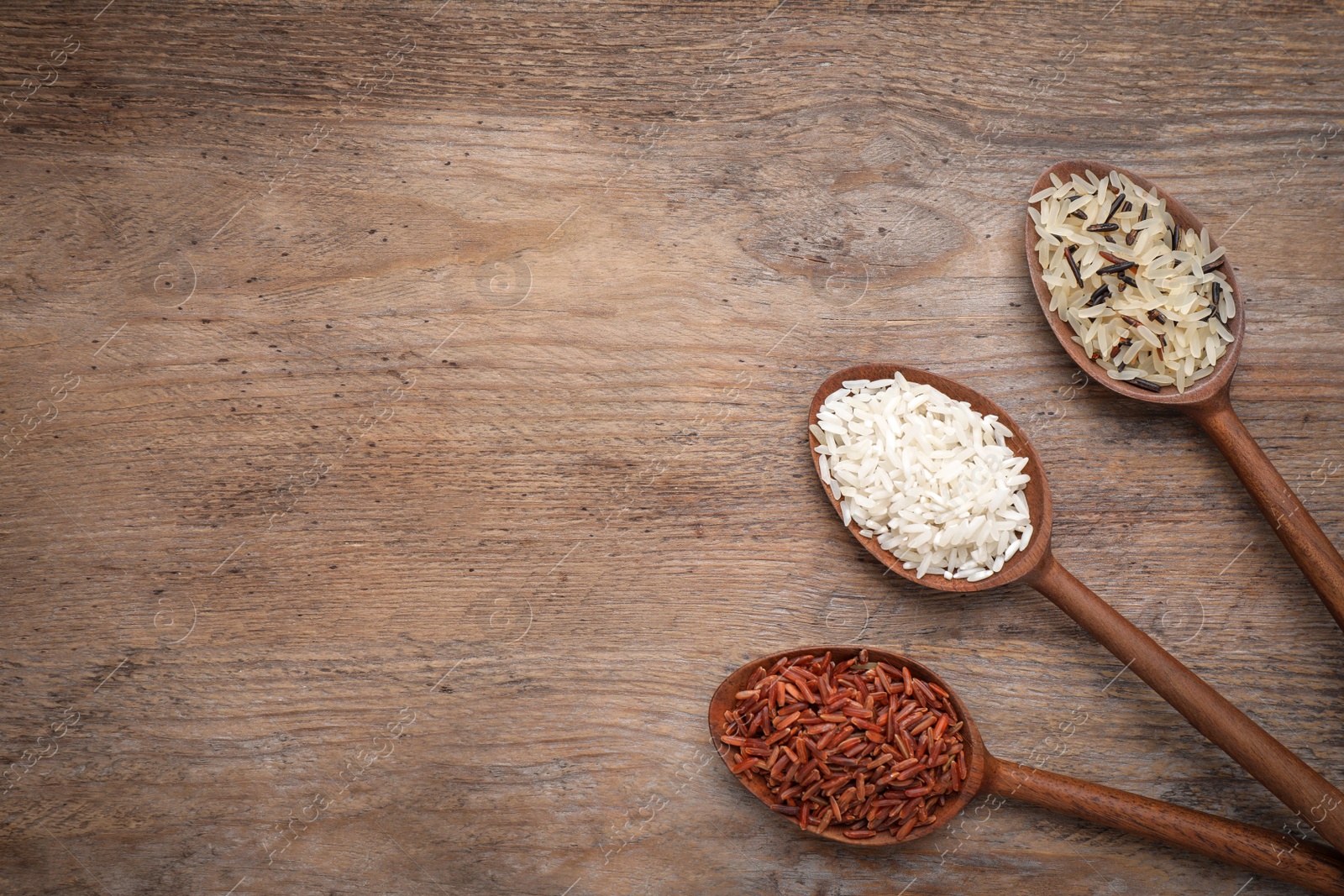 Photo of Brown and polished rice in spoons on wooden table, flat lay. Space for text