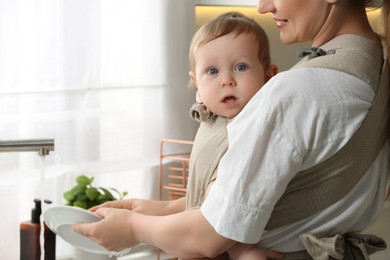 Mother holding her child in sling (baby carrier) while washing plates at home