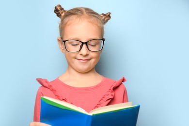 Cute little girl in glasses reading textbook on light blue background. Space for text