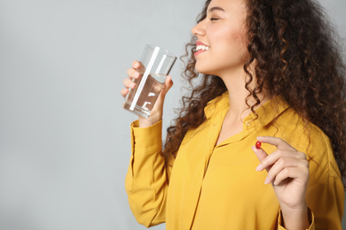 African-American woman with glass of water and vitamin capsule on light grey background