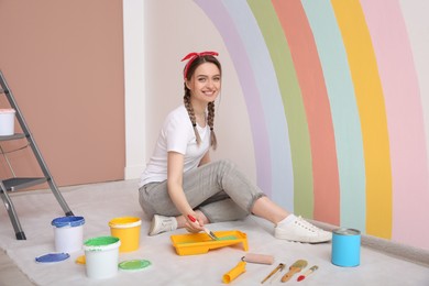 Photo of Young woman painting rainbow on white wall indoors