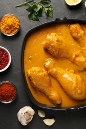 Tasty chicken curry and ingredients on black textured table, flat lay